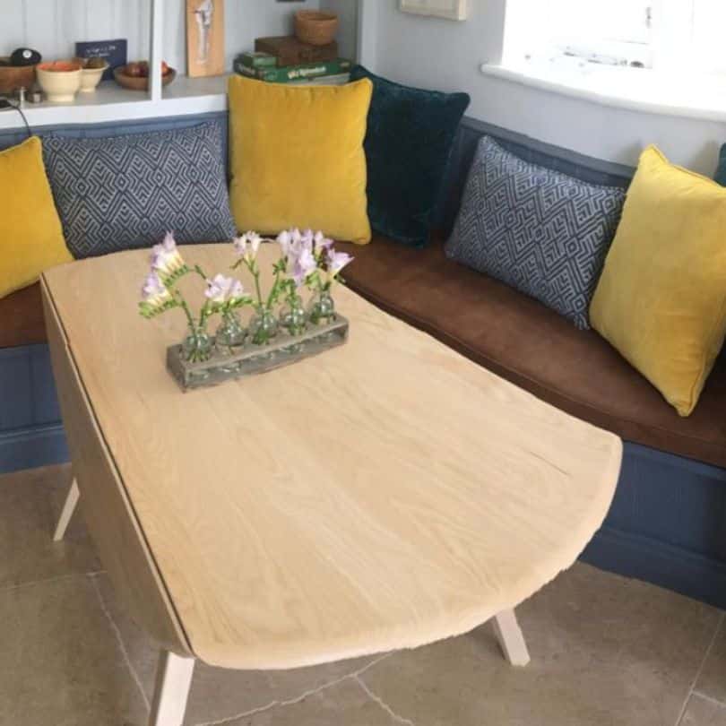 Banquette seating in brown fabric with grey and mustard cushions. Drop leaf table in pale wood with a flower arrangement